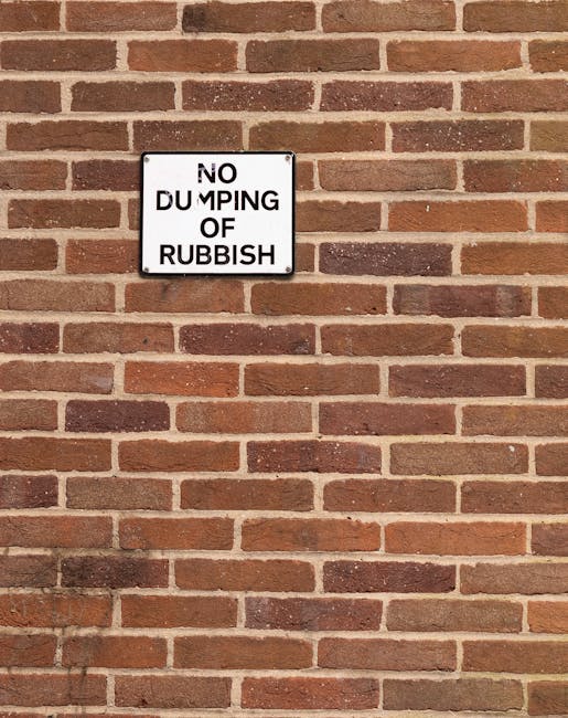 A close-up photograph of a brick wall with reddish-brown and darker brick tones arranged in a horizontal pattern. Mounted on the wall is a rectangular metal sign with a white background and black text that reads 'NO DUMPING OF RUBBISH', with the word 'DUMPING' partially obscured by wear or damage. The sign is positioned slightly above the center of the image, with the wall extending evenly on all sides. The texture of the bricks is rough with visible mortar joints, and the lighting is natural and diffuse, casting subtle shadows that highlight the brick surface. The context suggests a private or commercial property area where waste disposal is regulated, possibly part of a rubbish removal or clearance service area, as indicated by the company's focus on alternative waste handling.