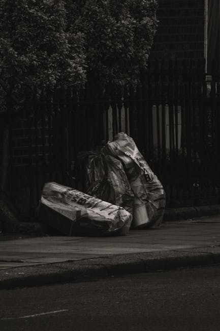 Two large black plastic refuse bags filled with waste materials are placed on the pavement in front of a black metal fence. The bags appear crumpled with visible creases and torn areas, indicating they are filled with unknown rubbish. The bags are positioned slightly leaning against each other, with one partially overlapping the other, and are situated at the edge of a residential street. Behind the fence, dense foliage from a tree or shrub partially obscures a building structure with horizontal wooden slats. The lighting suggests an overcast or shaded environment, with minimal natural light illuminating the scene. This scene depicts typical private waste disposal, often managed through independent rubbish collection services such as Waste Clearance Brompton, rather than municipal waste collection, supporting the context of alternative waste handling methods in Brompton.