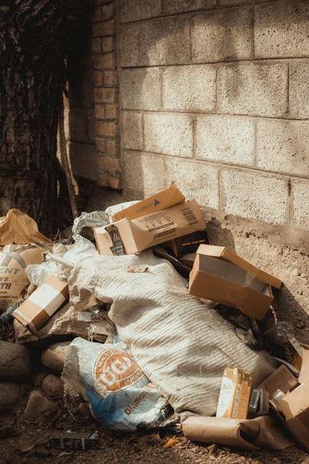 A close-up photograph of a brick wall with reddish-brown and darker brick tones arranged in a horizontal pattern. Mounted on the wall is a rectangular metal sign with a white background and black text that reads 'NO DUMPING OF RUBBISH', with the word 'DUMPING' partially obscured by wear or damage. The sign is positioned slightly above the center of the image, with the wall extending evenly on all sides. The texture of the bricks is rough with visible mortar joints, and the lighting is natural and diffuse, casting subtle shadows that highlight the brick surface. The context suggests a private or commercial property area where waste disposal is regulated, possibly part of a rubbish removal or clearance service area, as indicated by the company's focus on alternative waste handling.