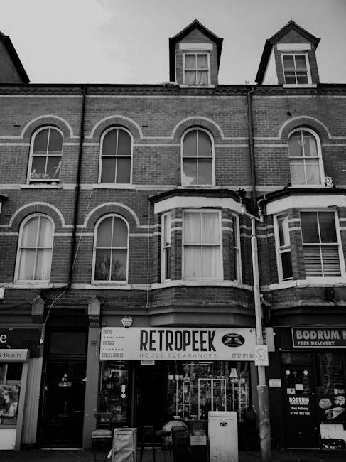 The image depicts a multi-story brick building with architectural details including arched windows on the upper floors and dormer windows on the roof. The ground floor features a shop with a large storefront window and a sign reading 'RETROPEEK' indicating house clearance services, which suggests a connection to private rubbish removal or alternative waste handling. The storefront is positioned centrally, with a small pavement area in front, containing a litter bin and some bags or containers placed outside.Adjacent to the 'RETROPEEK' shop, there is another storefront to the right with signage for a delivery service. The building's exterior appears aged, with visible brickwork and window frames. The overall scene is in black and white, emphasizing the textures of the brickwork and shop facades. The street setting, along with the clear signage, illustrates a typical commercial property engaged in household clearance or rubbish management activities, relevant to independent waste collection solutions services provided by Waste Clearance Brompton.