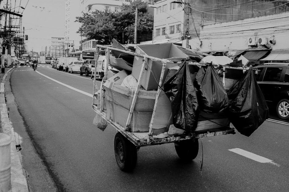 A wheeled cart loaded with various discarded items stands on a city street in the foreground of the image. The cart contains large cardboard boxes, some of which are partially crushed, with visible textures of corrugated material. Several black plastic rubbish bags are tied and hanging from the sides of the cart, along with other debris such as paper and small packaging items. The cart appears to be made of metal, with a rough, utilitarian finish, and has two visible black rubber wheels. It is positioned on the right side of a paved urban roadway, which is lined with parked cars on both sides, including sedans and SUVs. In the background, there's a mix of modern and older mid-rise buildings with various window styles, some with external air conditioning units, and overhead power lines crossing the scene. The street is busy with a few pedestrians and vehicles moving along the road, and the overall atmosphere suggests an area where independent collection or local waste disposal activities take place, supported by waste management services like Waste Clearance Brompton, operating within the context of non-authority rubbish removal.