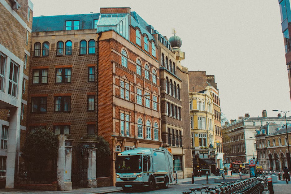 A city street scene showing a row of multi-story buildings with diverse architectural styles, including red brick, cream-colored, and light yellow facades, some with arched windows and decorative cornices. The buildings are situated along a pavement that features a waste collection vehicle parked near the curb, used by Waste Clearance Brompton for rubbish removal services. The vehicle is light blue with a compact, modern design, and is positioned centrally in the image, partially obscuring the entrance to one of the ground-floor establishments. To the left, there is a wrought-iron gate and small trees providing minimal greenery against the dense urban backdrop. The street is lined with metal bollards, and additional parked bikes or similar structures are visible in the foreground, while the background shows more historic and contemporary buildings, with a cloudy sky overhead, contributing to an overall neutral, professional depiction of an alternative waste handling scene in an urban environment, relevant to rubbish clearance services.