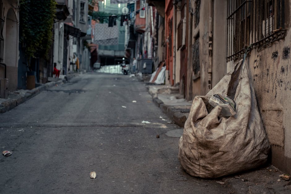 A wheeled cart loaded with various discarded items stands on a city street in the foreground of the image. The cart contains large cardboard boxes, some of which are partially crushed, with visible textures of corrugated material. Several black plastic rubbish bags are tied and hanging from the sides of the cart, along with other debris such as paper and small packaging items. The cart appears to be made of metal, with a rough, utilitarian finish, and has two visible black rubber wheels. It is positioned on the right side of a paved urban roadway, which is lined with parked cars on both sides, including sedans and SUVs. In the background, there's a mix of modern and older mid-rise buildings with various window styles, some with external air conditioning units, and overhead power lines crossing the scene. The street is busy with a few pedestrians and vehicles moving along the road, and the overall atmosphere suggests an area where independent collection or local waste disposal activities take place, supported by waste management services like Waste Clearance Brompton, operating within the context of non-authority rubbish removal.