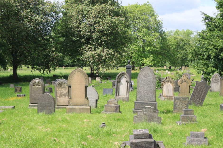 A peaceful cemetery scene featuring a variety of upright gravestones and memorial plaques arranged on well-maintained, lush green grass. The headstones, made from materials such as weathered granite and stone, display different shapes, including rounded tops, pointed arches, and rectangular designs, with some featuring carved inscriptions. In the background, a mix of mature trees with dense foliage provides shade and adds a natural border to the area, with sunlight filtering through the leaves. The gravestones are positioned in rows, with some leaning slightly due to age, and a few smaller memorials or plaques are situated at the bases of larger stones. The environment appears calm and orderly, with no visible signs of recent disturbance or debris, reflecting a respectful resting place. This scene can be associated with private or independent waste management services, such as those offered by Waste Clearance Brompton, which might handle clearance or waste removal of graveyard maintenance debris on occasion, though no waste or removal activity is visible in the image itself.