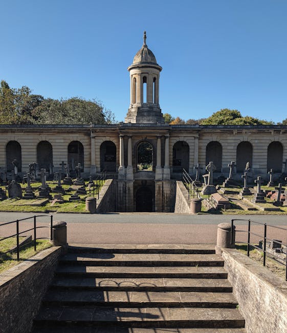 A peaceful cemetery scene featuring a variety of upright gravestones and memorial plaques arranged on well-maintained, lush green grass. The headstones, made from materials such as weathered granite and stone, display different shapes, including rounded tops, pointed arches, and rectangular designs, with some featuring carved inscriptions. In the background, a mix of mature trees with dense foliage provides shade and adds a natural border to the area, with sunlight filtering through the leaves. The gravestones are positioned in rows, with some leaning slightly due to age, and a few smaller memorials or plaques are situated at the bases of larger stones. The environment appears calm and orderly, with no visible signs of recent disturbance or debris, reflecting a respectful resting place. This scene can be associated with private or independent waste management services, such as those offered by Waste Clearance Brompton, which might handle clearance or waste removal of graveyard maintenance debris on occasion, though no waste or removal activity is visible in the image itself.