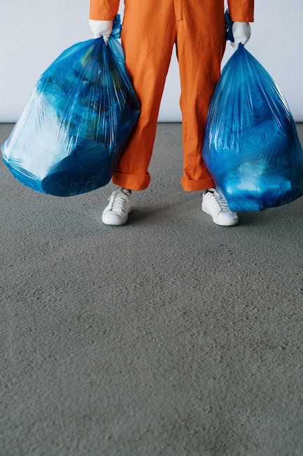 A person wearing orange work overalls and white sneakers is standing on a grey carpeted floor, holding two large blue rubbish bags filled with waste, one in each hand, with the bags appearing to be made of transparent plastic. The background shows a plain white wall, and the scene suggests a waste collection or rubbish removal activity, possibly related to private or commercial waste handling services such as those offered by Waste Clearance Brompton. The focus is on the individual's lower body and the filled rubbish bags, which are slightly leaning and bulging with waste material, indicating an active rubbish collection process in an indoor or enclosed environment. The overall setting emphasizes the practical aspect of rubbish disposal, aligning with contexts of independent waste clearance or on-site rubbish removal, with no additional objects or tools visible in the frame.