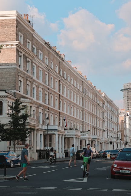 A wide view of a street scene in front of a row of elegant, multi-storey Victorian-style terraced houses with light-colored stone facades, decorative cornices, and sash windows, set against a partly cloudy blue sky. In the foreground, pedestrians are crossing the street, including a man on a bicycle with a green frame and a woman walking along the pavement. Several parked cars line the street, including a red car visible on the right side. A small tree is planted along the sidewalk, and street lamps are mounted on the building facades near the entrances. One building appears to be representative of private rental or disposal options, suggesting a context for alternative waste handling or rubbish removal services suited for residential properties, as offered by Waste Clearance Brompton. In the background, a construction site with scaffolding indicates ongoing development or renovation in the area, contributing to the urban environment's character. The scene is well-lit with natural daylight, emphasizing the clean facade of the housing and the casual, everyday activity of city life, relevant to professional rubbish removal and waste clearance services in South Kensington and Brompton areas.