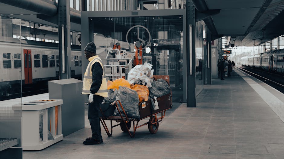 A person wearing orange work overalls and white sneakers is standing on a grey carpeted floor, holding two large blue rubbish bags filled with waste, one in each hand, with the bags appearing to be made of transparent plastic. The background shows a plain white wall, and the scene suggests a waste collection or rubbish removal activity, possibly related to private or commercial waste handling services such as those offered by Waste Clearance Brompton. The focus is on the individual's lower body and the filled rubbish bags, which are slightly leaning and bulging with waste material, indicating an active rubbish collection process in an indoor or enclosed environment. The overall setting emphasizes the practical aspect of rubbish disposal, aligning with contexts of independent waste clearance or on-site rubbish removal, with no additional objects or tools visible in the frame.